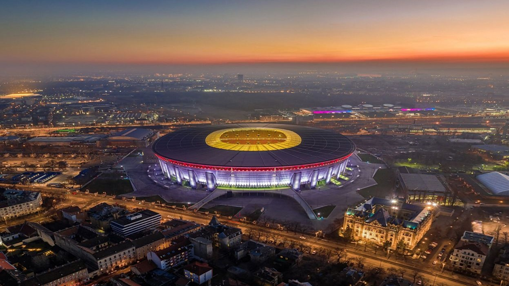 Légi felvétel egy kivilágított stadionról naplementekor, városi utcákkal és épületekkel körülötte.